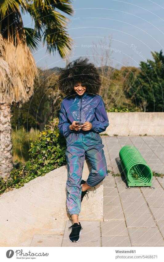 Stylish young woman wearing tracksuit and using cell phone outdoors human human being human beings humans person persons curl curled curls curly hair