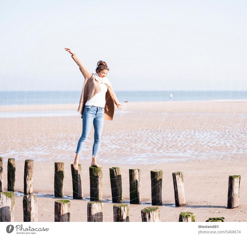 Smiling young woman with arms outstretched walking on wooden posts at beach during sunny day color image colour image Netherlands Holland The Netherlands