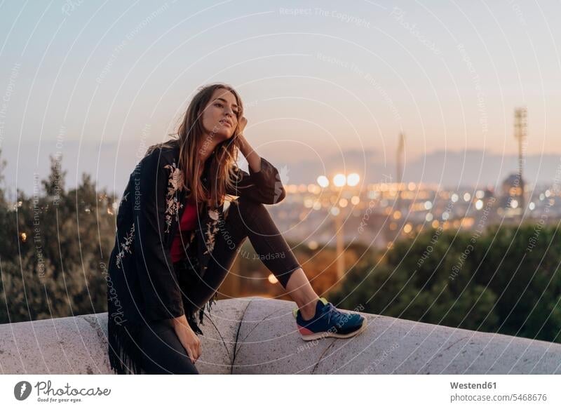Spain, Barcelona, Montjuic, young woman sitting on a wall at dusk with city lights in background walls females women atmosphere atmospheric evening