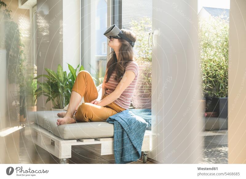 Young woman sitting at the window at home wearing VR glasses windows females women virtual Seated specs Eye Glasses spectacles Eyeglasses Adults grown-ups