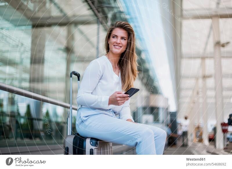 Smiling young businesswoman sitting on luggage with cell phone looking around Seated looking round look round look around baggage mobile phone mobiles