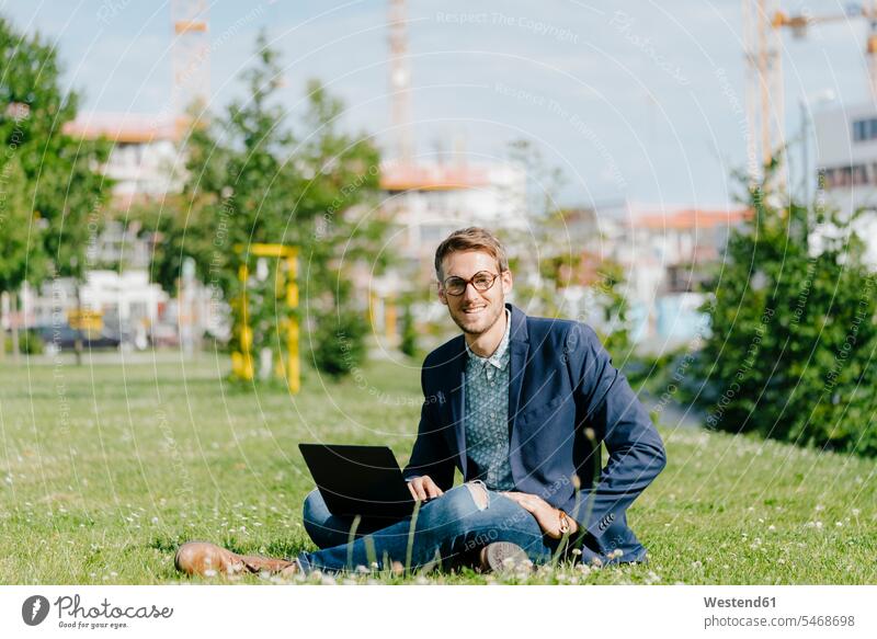 Young businessman sitting in park, using laptop grass Grassy parks cross-legged tailor seat young man young men Laptop Computers laptops notebook Businessman