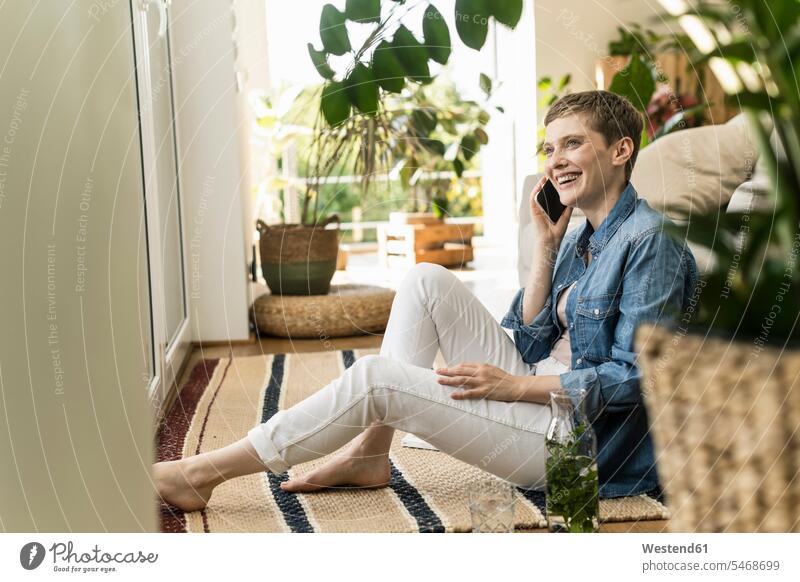 Cheerful woman talking over smart phone while sitting on carpet at home color image colour image Germany leisure activity leisure activities free time