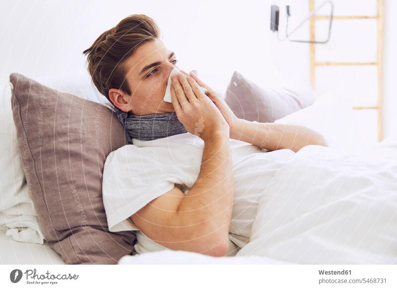 Sick young man blowing nose while lying on bed at home color image colour image Germany indoors indoor shot indoor shots interior interior view Interiors