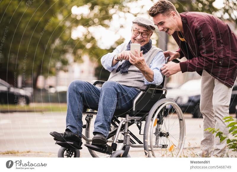 Laughing senior man in wheelchair and his adult grandson looking together at smartphone having fun human human being human beings humans person persons