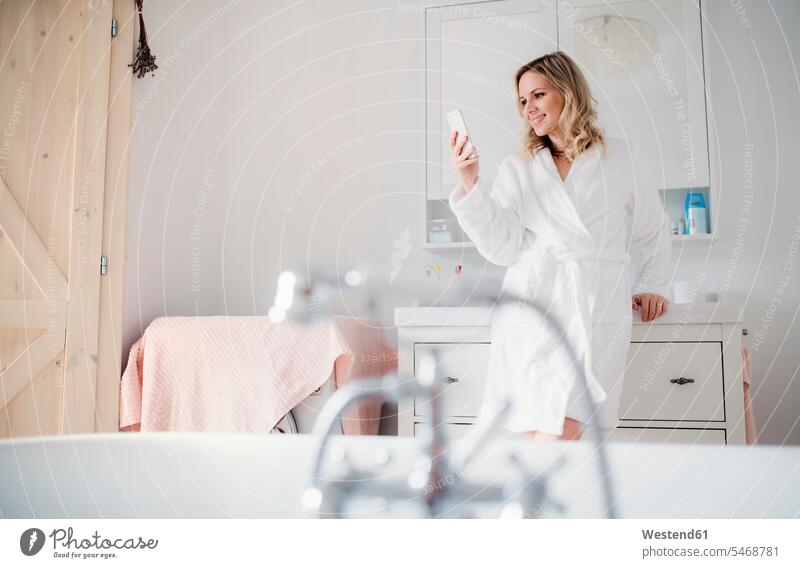 Smiling woman wearing bathrobe in bathroom at home holding cell phone females women morning in the morning smiling smile mobile phone mobiles mobile phones