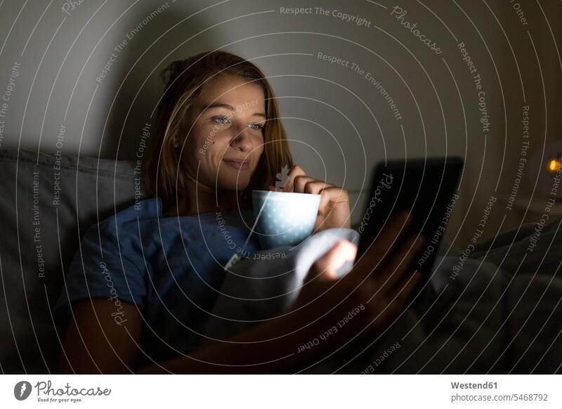 Young woman lying in bed at home with cup of tea at night using tablet human human being human beings humans person persons caucasian appearance