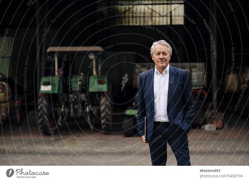 Portrait of senior businessman holding laptop on a farm with tractor in barn Occupation Work job jobs profession professional occupation business life
