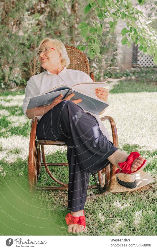 Senior woman holding book looking away while relaxing on chair in yard color image colour image Spain leisure activity leisure activities free time leisure time