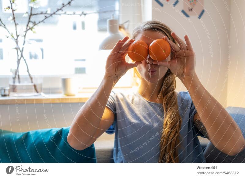Playful young woman covering her eyes with oranges at home human human being human beings humans person persons celibate celibates singles solitary people