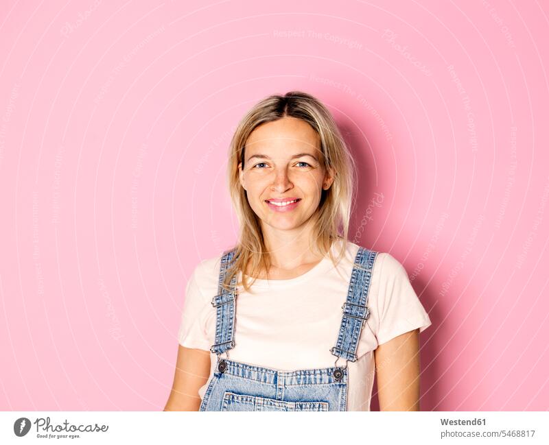 Portrait of smiling blond woman wearing dungarees in front of pink background blond hair blonde hair smile Bib Overall Bibs Overall Bib Overalls portrait