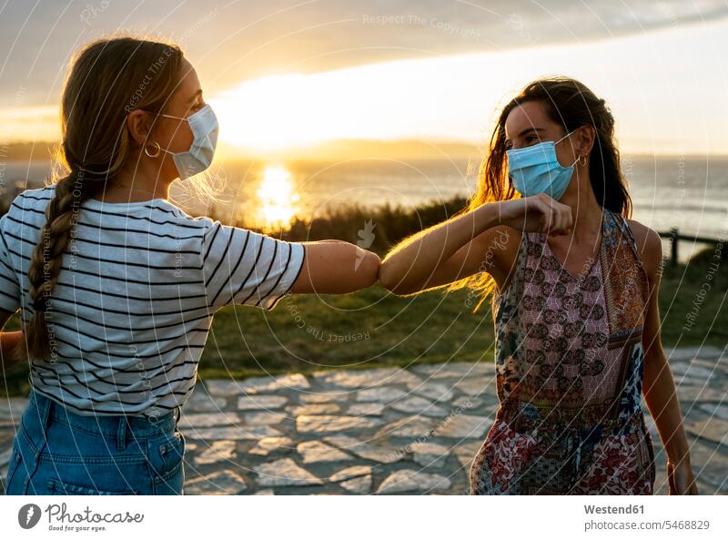 Women in protective face masks elbow bumping against sky during COVID-19 outbreak color image colour image outdoors location shots outdoor shot outdoor shots