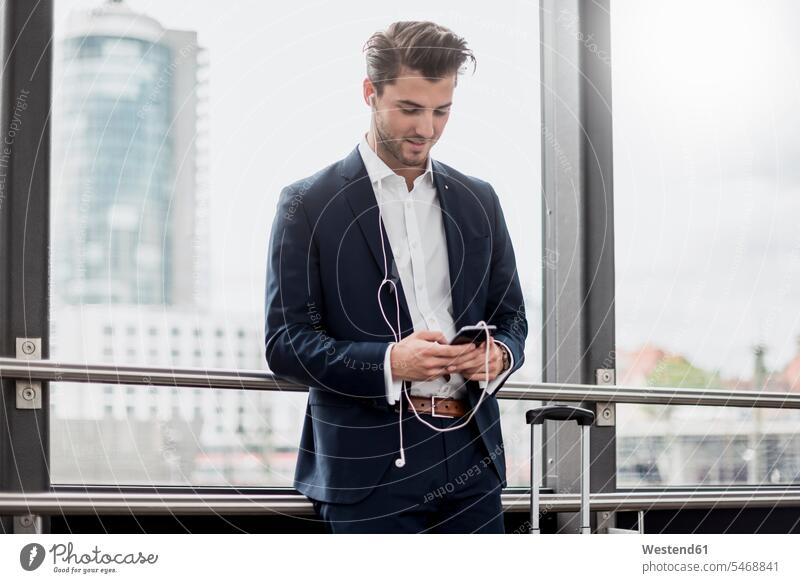Young businessman at the window with cell phone, earbuds and rolling suitcase Businessman Business man Businessmen Business men earphones ear phone ear phones
