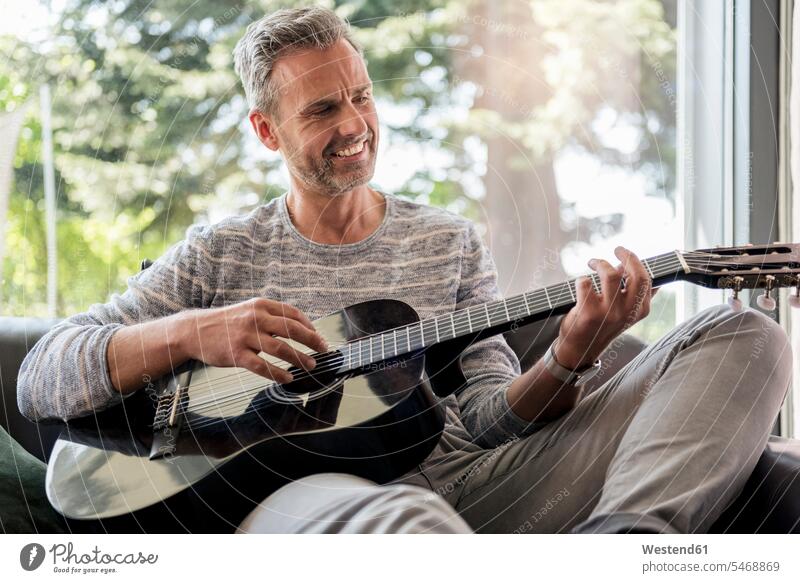 Happy mature man lying on couch at home playing guitar guitars smiling smile sitting Seated settee sofa sofas couches settees men males happiness happy