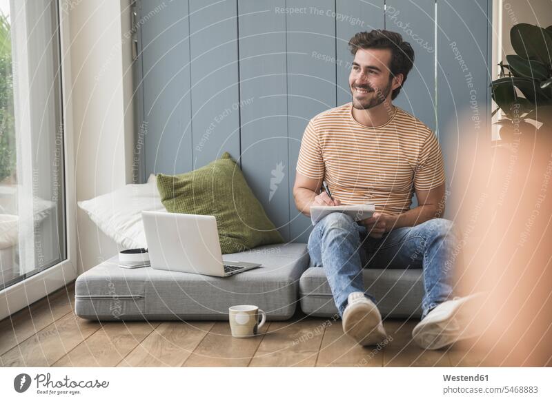 Young man sitting on mattress, using laptop, taking notes Germany notepad pads Note Pad notepads Note Pads toothy smile big smile open smile laughing wireless