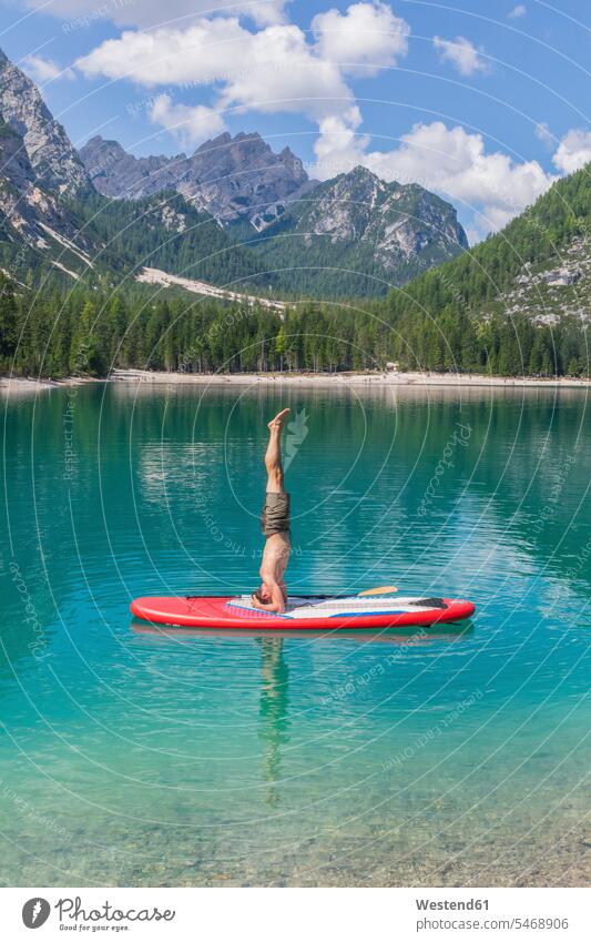 Man doing headstand on paddleboard at Pragser Wildsee against mountain range, Dolomites, Alto Adige, Italy color image colour image outdoors location shots