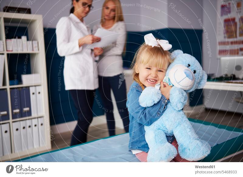 Girl hugging teddy in medical practice with doctor and mother in background teddies girl females girls Female Doctor physicians Female Doctors mommy mothers