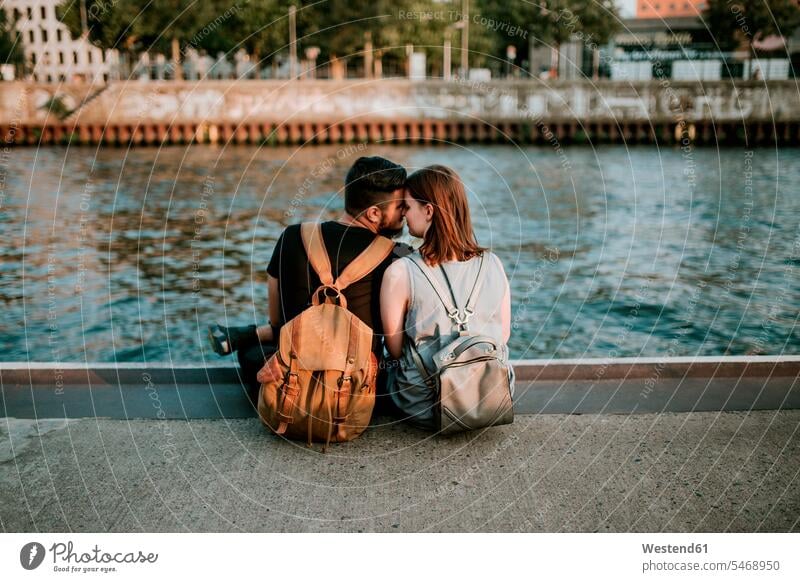 Affectionate young couple sitting at River Spree, Berlin, Germany touristic tourists back-pack back-packs backpacks rucksack rucksacks cuddle snuggle snuggling