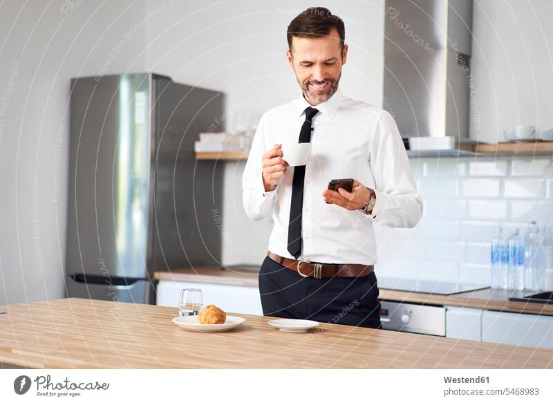 Man standing int he kitchen in the having coffee and looking at his phone before work drinking using use Smartphone iPhone Smartphones morning in the morning