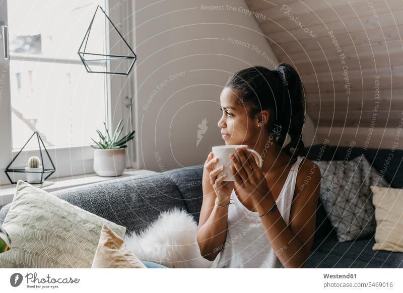 Young woman with cup of coffee sitting on the couch at home looking out of window Coffee Cup Coffee Cups females women Seated windows settee sofa sofas couches