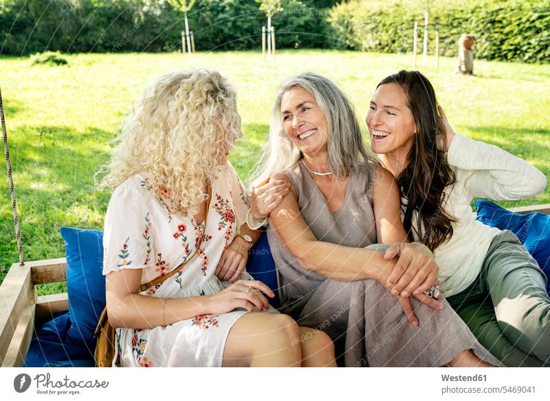 Three happy women of different age sittingon a hanging bed in garden talking speaking relaxed relaxation Seated happiness woman females age difference