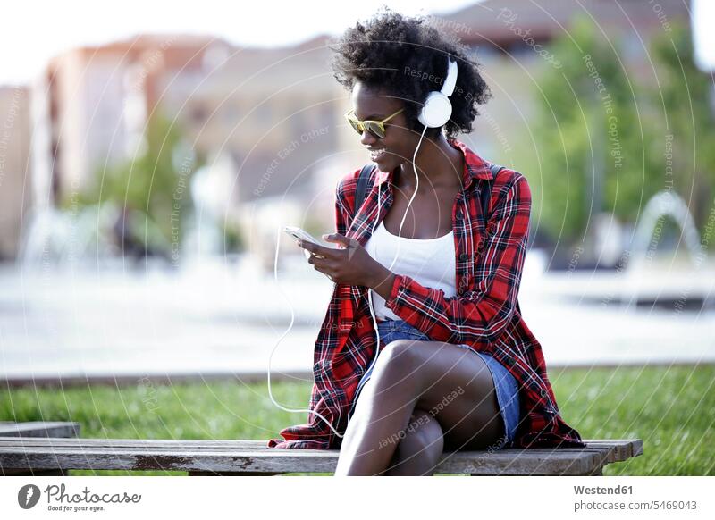 Smiling young woman sitting on bench in city park listening music with headphones Smartphone iPhone Smartphones hearing Seated smiling smile females women