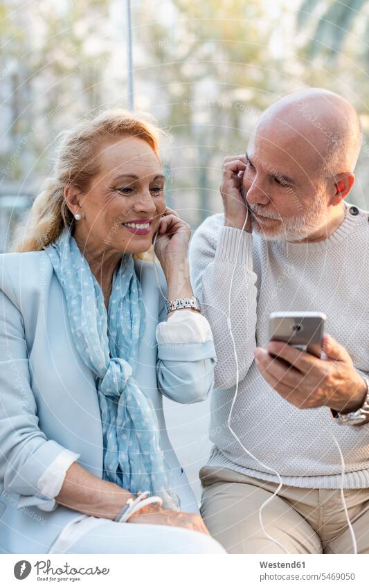 Spain, Barcelona, happy senior couple sharing smartphone with earbuds earphones ear phone ear phones share twosomes partnership couples happiness Smartphone