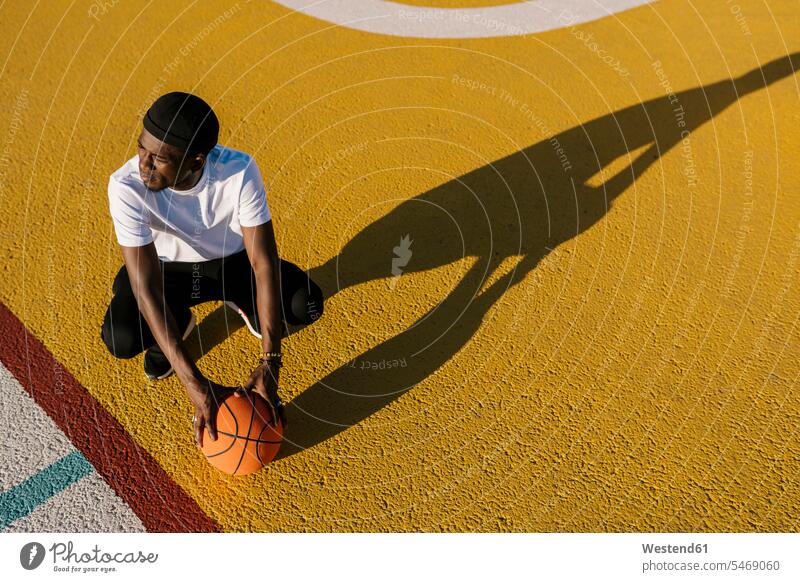 Thoughtful young man holding basketball crouching on sports court during sunny day color image colour image Spain leisure activity leisure activities free time