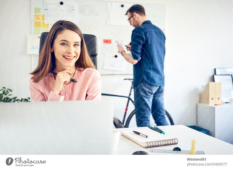 Portrait of smiling woman working at desk in office with colleague in background desks colleagues At Work portrait portraits smile businesswoman businesswomen