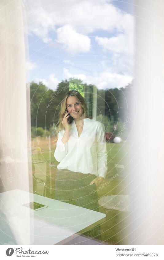 Smiling businesswoman talking on the phone at home behind windowpane human human being human beings humans person persons caucasian appearance