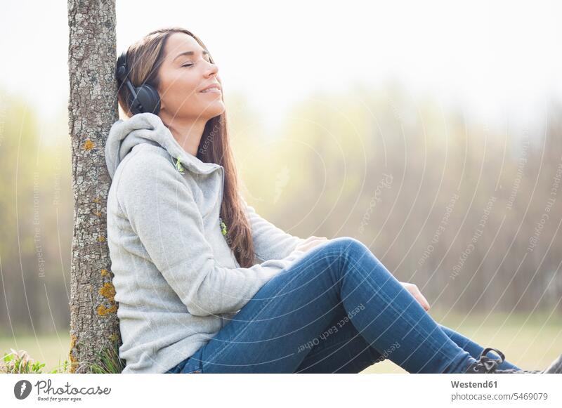 Relaxed woman listening music while leaning on tree trunk at park color image colour image outdoors location shots outdoor shot outdoor shots day daylight shot