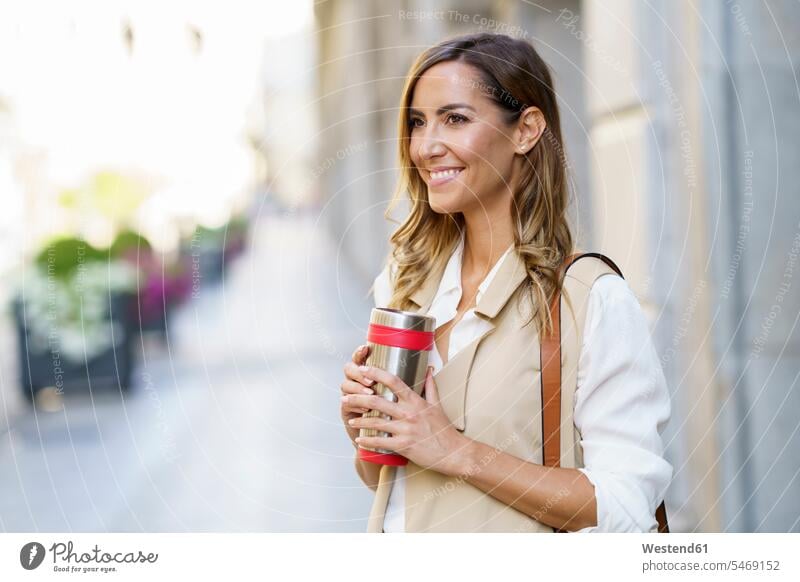 Smiling woman holding metal thermos while standing at sidewalk in city color image colour image outdoors location shots outdoor shot outdoor shots day