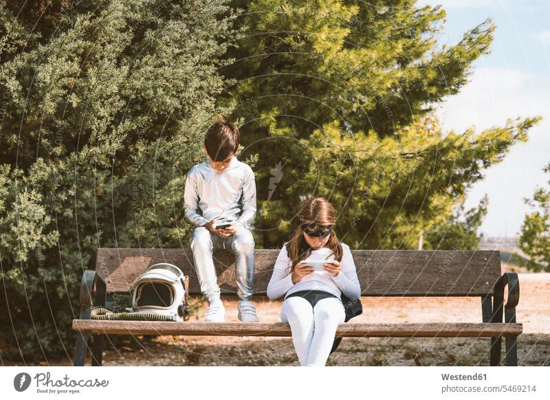 Two kids in astronaut and superhero costumes using mobile phone on park bench friends mate human human being human beings humans person persons braver bravers