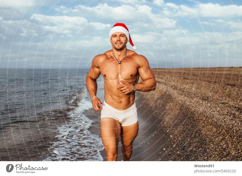 Smiling muscular young man wearing Santa hat running at beach against cloudy sky color image colour image Spain leisure activity leisure activities free time