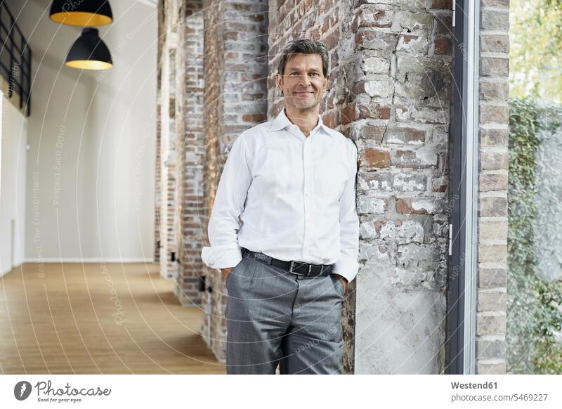Portrait of smiling businessman leaning against brick wall in modern office offices office room office rooms portrait portraits brick walls smile contemporary