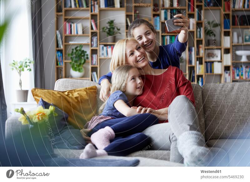 Mother and her daughters sitting on couch,taking smartphone selfies Seated settee sofa sofas couches settees mother mommy mothers mummy mama photographing