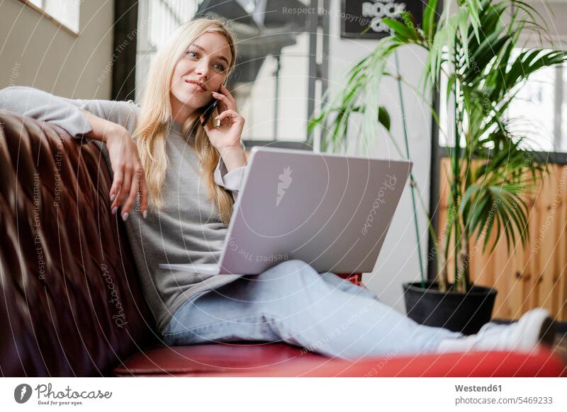 Smiling young woman sitting on couch with laptop talking on cell phone females women on the phone call telephoning On The Telephone calling Seated smiling smile