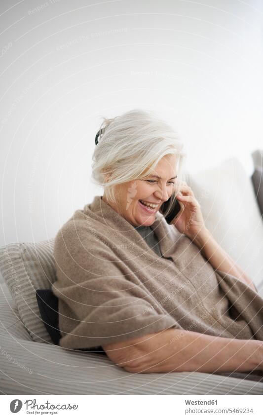 Cheerful senior woman talking on mobile phone while sitting against white wall at home color image colour image Denmark Scandinavia Scandinavian Peninsula