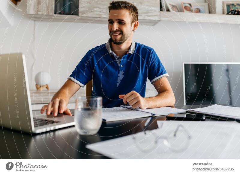 Smiling handsome young man using laptop while doing homework at table color image colour image indoors indoor shot indoor shots interior interior view Interiors