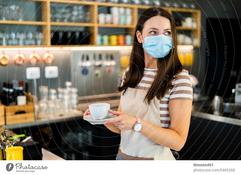 Thoughtful young owner wearing mask while holding coffee cup and saucer in cafe color image colour image indoors indoor shot indoor shots interior interior view