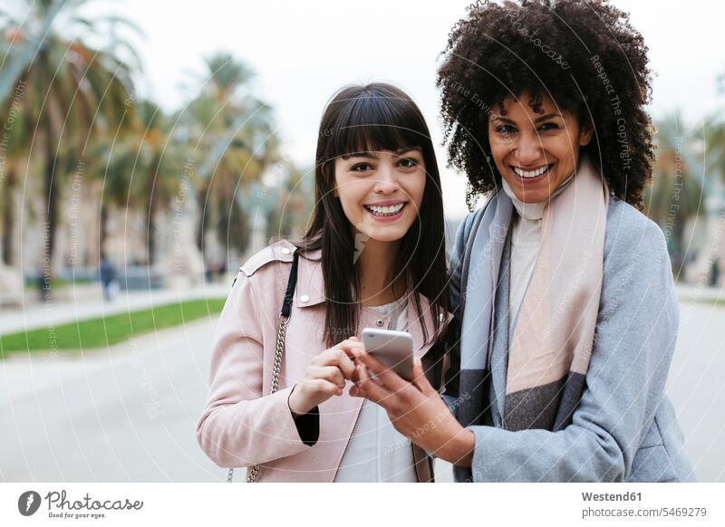 Spain, Barcelona, portrait of two happy women with cell phone on promenade portraits promenades female friends mobile phone mobiles mobile phones Cellphone