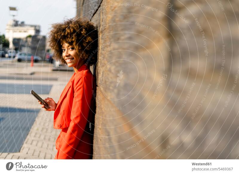 Portrait of smiling young woman with cell phone wearing fashionable red pantsuit leaning against wall telecommunication phones telephone telephones cell phones