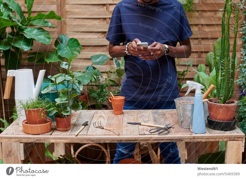 Young man using smartphone on his terrace while gardening gardeners flower pot flower pots flowerpots Tables wood wood table telecommunication phones telephone