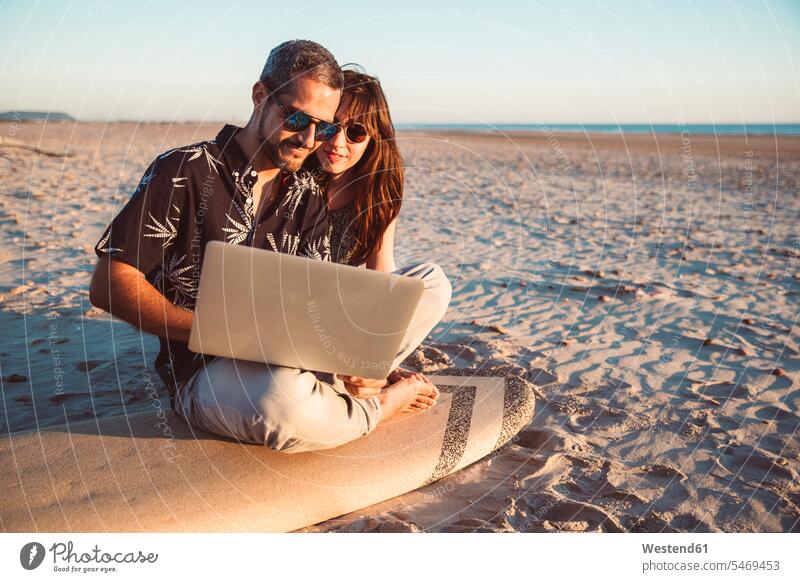 Couple with surfboard sitting on the beach, using laptop couple twosomes partnership couples beaches surfboards Laptop Computers laptops notebook using a laptop