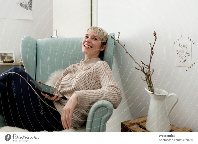 Happy woman sitting in armchair at home with tablet Germany resting flat flats apartment apartments jar jugs jars toothy smile big smile open smile laughing
