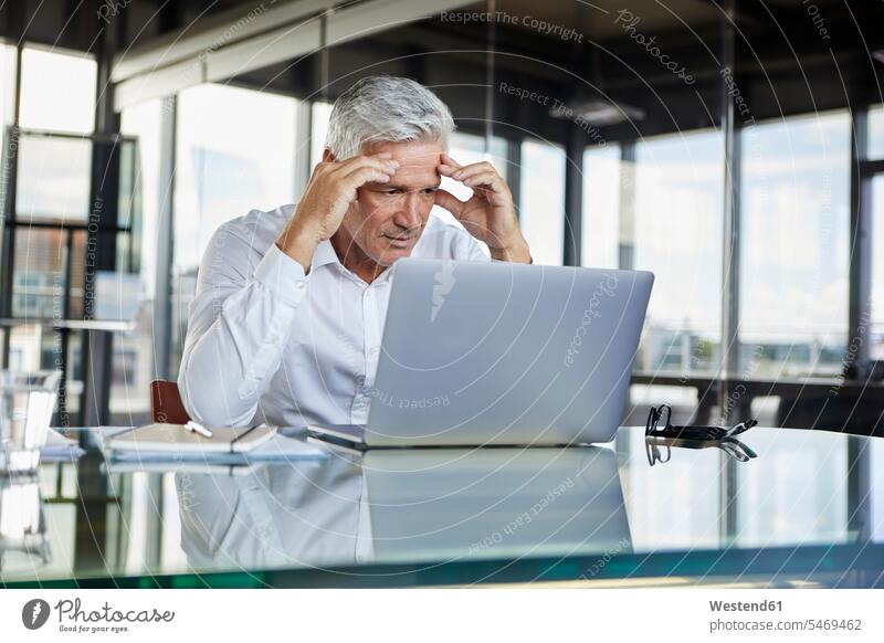 Stressed businessman sitting at desk with laptop, holding his head Office Offices Businessman Business man Businessmen Business men desks using laptop