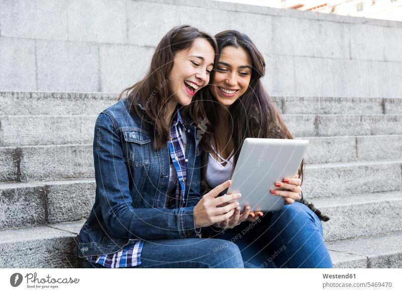 Happy young women watching tablet sitting on steps together in Madrid, Spain human human being human beings humans person persons caucasian appearance