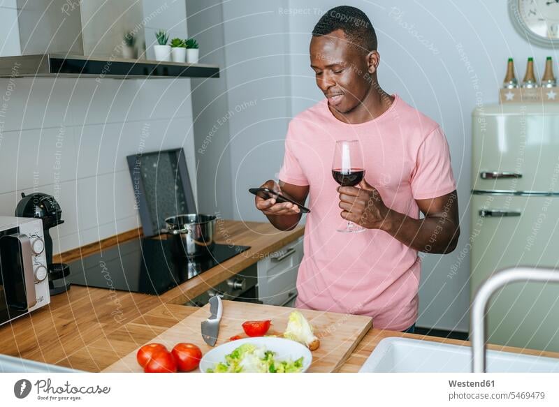Young man with wine glass using cell phone while preparing salad human human being human beings humans person persons African black black ethnicity coloured 1