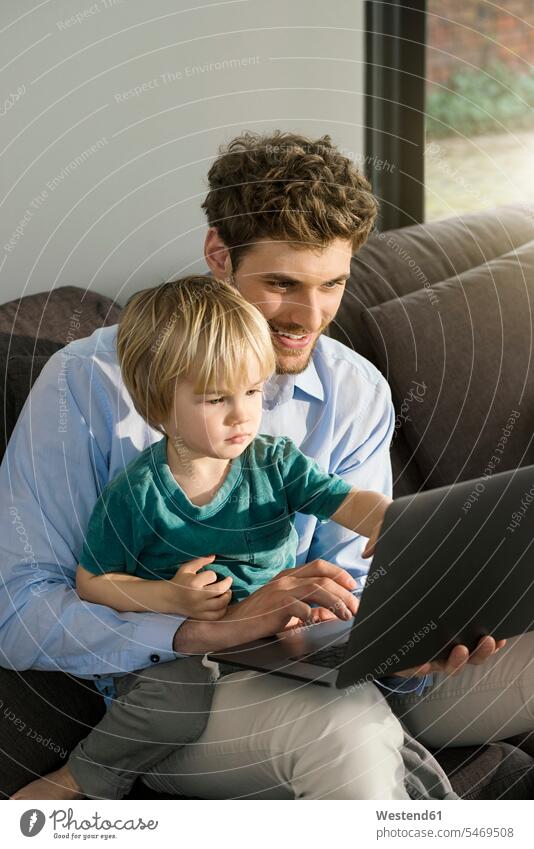 Father and son looking at laptop on couch at home Laptop Computers laptops notebook sitting Seated sons manchild manchildren settee sofa sofas couches settees