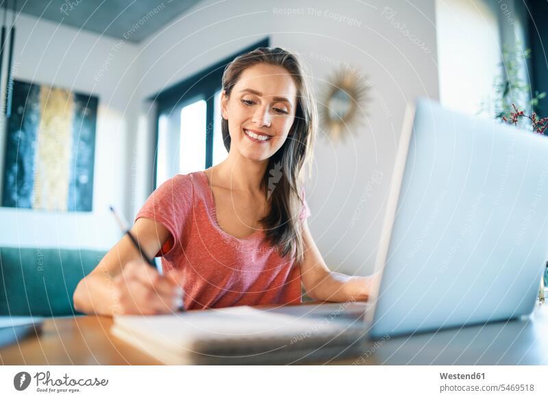 Smiling female freelancer writing in note pad on desk at home office color image colour image casual clothing casual wear leisure wear casual clothes